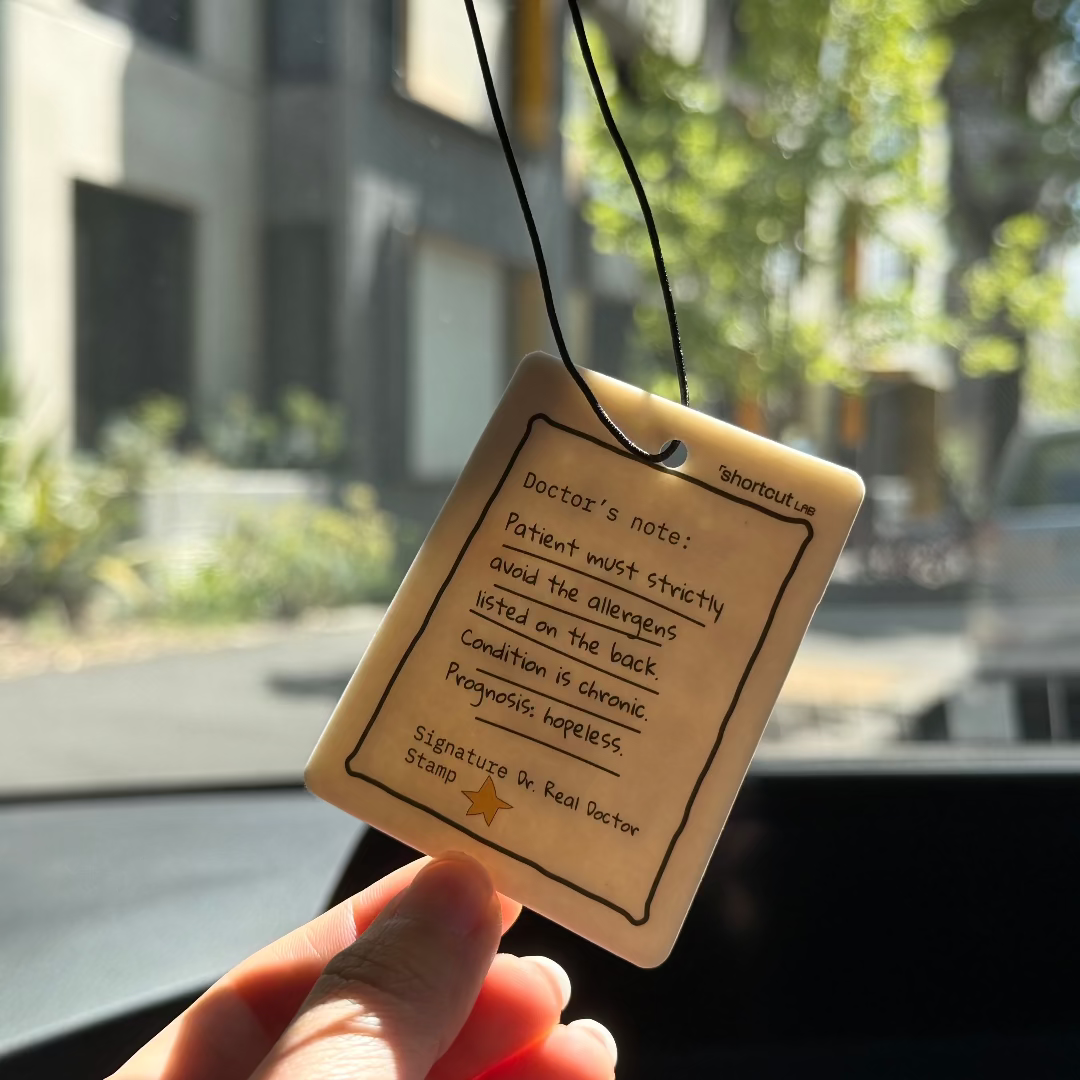 Shortcut Lab ‘Doctor’s Note’ car fragrance card in Amber Orchard scent featuring handwritten-style humour reading ‘Patient must strictly avoid the allergens listed on the back,’ hanging from a car rear-view mirror.