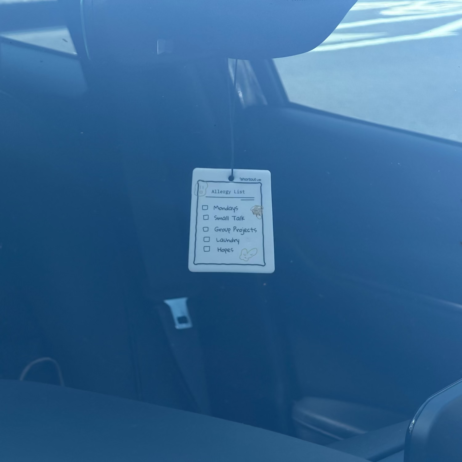 Shortcut Lab ‘Allergy List’ car fragrance card in Amber Orchard scent hanging from a rear-view mirror inside a car