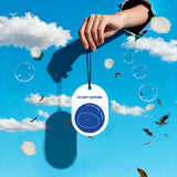 Shortcut Lab ‘Do Not Disturb’ car fragrance card in White Tea scent held against a blue sky with clouds and bubbles, featuring blue smiling face design and text ‘I’m busy doing nothing