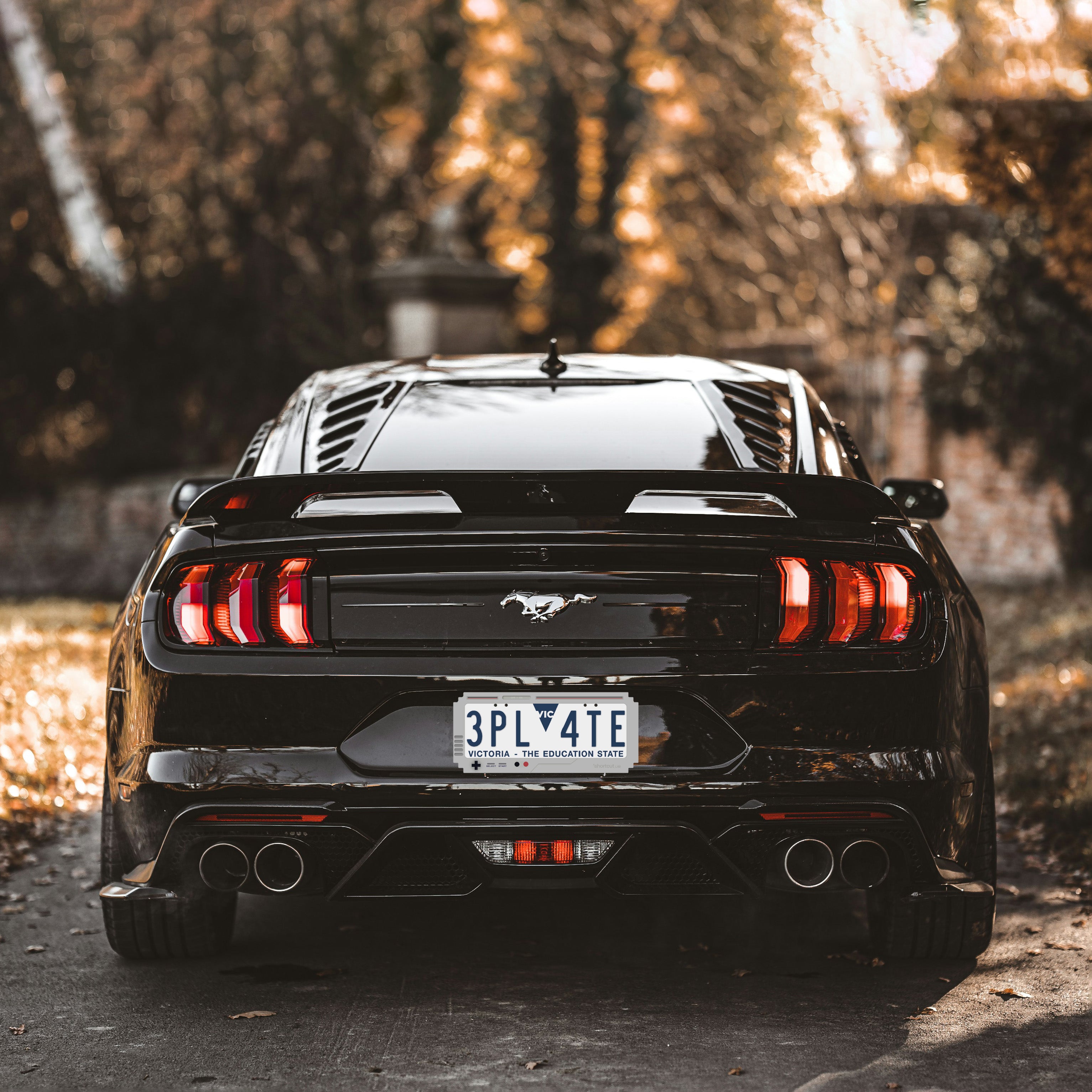 Shortcut Lab GameBoy-inspired number plate frame installed on a black sports car, rear view shot in warm sunlight surrounded by trees